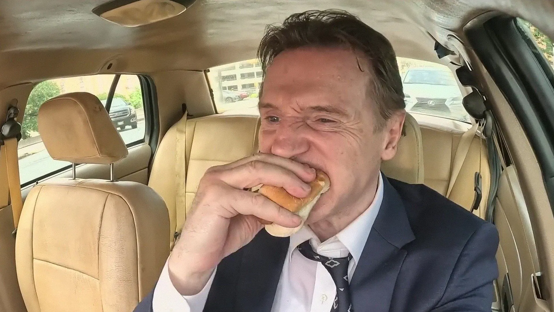 Movie Still from The Naked Gun 2025 Movie's Chili Dog scene. Features Lead Character in an old model Police car, eating a Chili Dog.