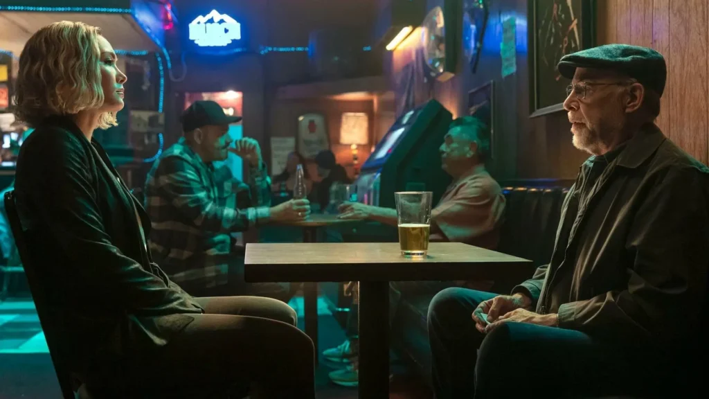 Movie still from The Accountant 2 - Raymond King (J.K. Simmons) in a diner booth, sitting across from Anaïs, in a dimly lit, blue-hued setting.