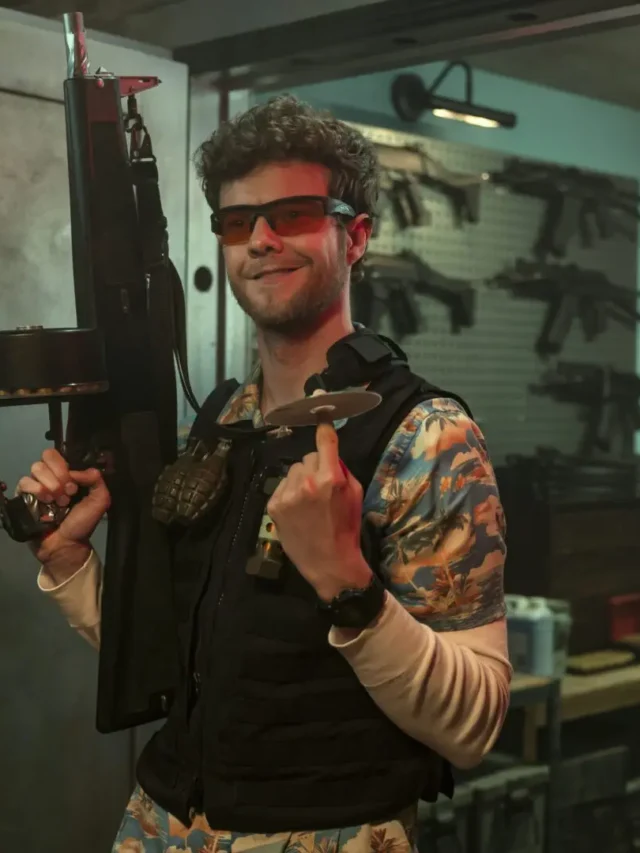 Jack Quaid wearing sunglasses and a Hawaiian shirt, holding a large firearm with a drum magazine, in a room full of weapons from Heads of State.